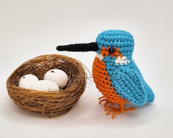 May include: A crocheted kingfisher bird with bright blue wings, an orange chest, and a long black beak, positioned next to a woven brown nest containing three small, smooth, white eggs with tiny black speckles on their surface.