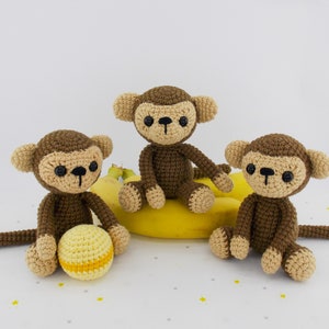 Può includere: Tre giocattoli di scimmia di crochet marroni seduti su una buccia di banana gialla. La scimmia a sinistra tiene una palla gialla.