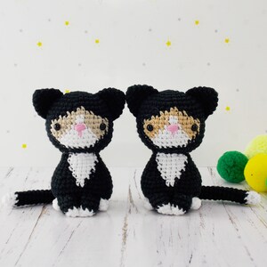 May include: Two crocheted black and white cat plushies with pink noses. The plushies are sitting on a white surface.