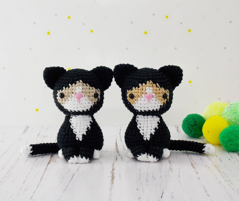 May include: Two crocheted black and white cat plushies with pink noses. The plushies are sitting on a white surface.