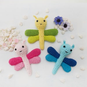 May include: Three crocheted dragonfly toys in different colors: pink, yellow, and blue. The toys have black eyes and are sitting on a white background.