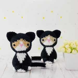 May include: Two crocheted black and white cat toys with orange accents. The cats have pink noses and black eyes. The cats are sitting on a white wooden surface.