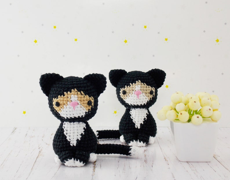 May include: Two crocheted black and white cat toys with orange accents. The cats have pink noses and black eyes. The cats are sitting on a white wooden surface.