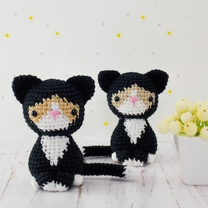 May include: Two black and white crocheted cat toys with orange accents. The cats are sitting on a white wooden surface.