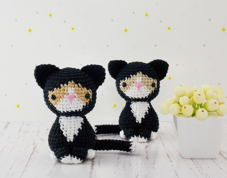 May include: Two black and white crocheted cat toys with orange accents. The cats are sitting on a white wooden surface.