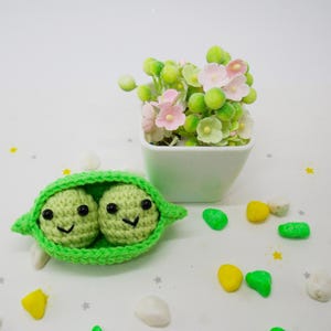 Pu&ograve; includere: Un baccello di piselli all'uncinetto fatto a mano con due piselli sorridenti. Il baccello &egrave; verde brillante e i piselli sono verde chiaro con occhi neri e sorrisi. Un piccolo vaso quadrato bianco con fiori rosa e verdi sullo sfondo.