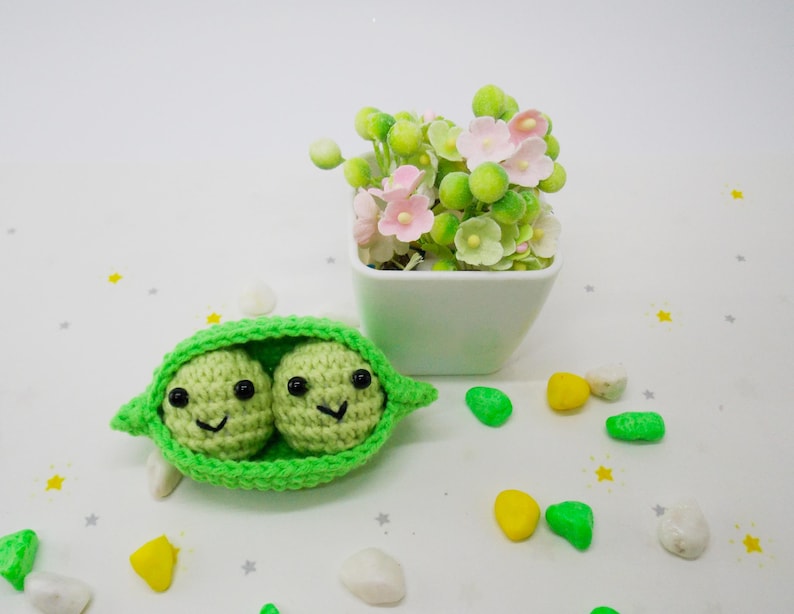 Pu&ograve; includere: Un baccello di piselli all'uncinetto fatto a mano con due piselli sorridenti. Il baccello &egrave; verde brillante e i piselli sono verde chiaro con occhi neri e sorrisi. Un piccolo vaso quadrato bianco con fiori rosa e verdi sullo sfondo.