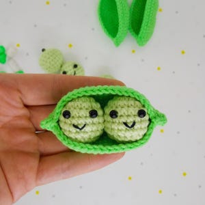 Pu&ograve; includere: Baccello di piselli all'uncinetto con due piselli sorridenti. Il baccello &egrave; verde brillante, con due piselli verde chiaro all'interno, ciascuno con occhi neri e un sorriso cucito. Altri piselli e foglie di baccello all'uncinetto sono visibili sullo sfondo.