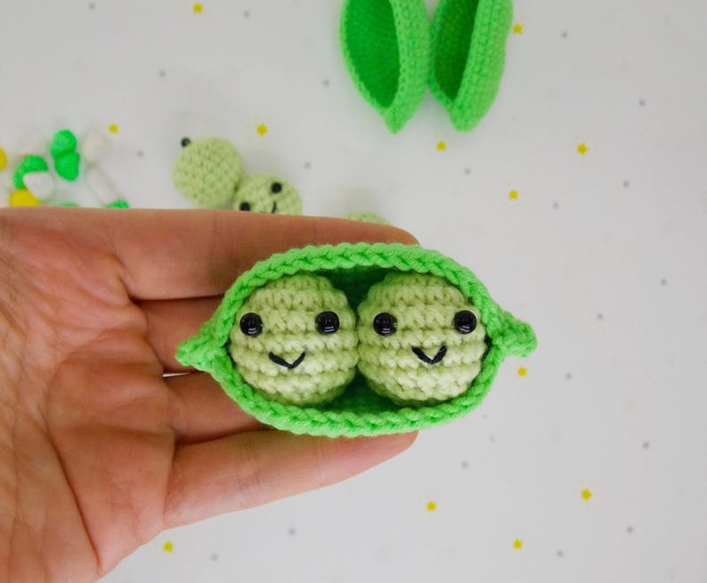 Pu&ograve; includere: Baccello di piselli all'uncinetto con due piselli sorridenti. Il baccello &egrave; verde brillante, con due piselli verde chiaro all'interno, ciascuno con occhi neri e un sorriso cucito. Altri piselli e foglie di baccello all'uncinetto sono visibili sullo sfondo.