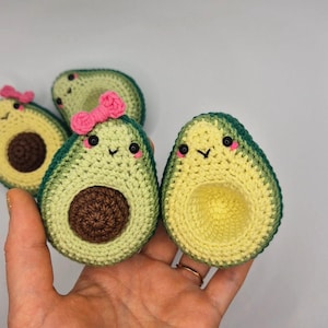 Puede incluir: Peluche de aguacate tejido a mano. Los aguacates son verdes y amarillos, con un borde verde oscuro. Un aguacate tiene un lazo rosa. Cada aguacate tiene una cara sonriente y un hueso marrón oscuro. Los peluches tienen un diseño caprichoso.