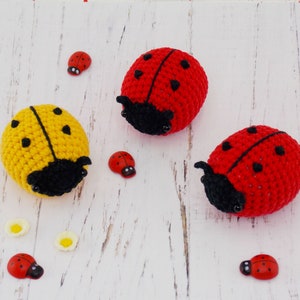 May include: Two crocheted ladybirds, one red and one yellow, on a white wooden background. There are several smaller red ladybirds scattered around the larger ones.