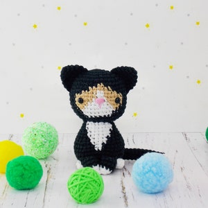 May include: A black and white crocheted cat toy with orange accents. The cat is sitting on a white surface with colorful balls of yarn.
