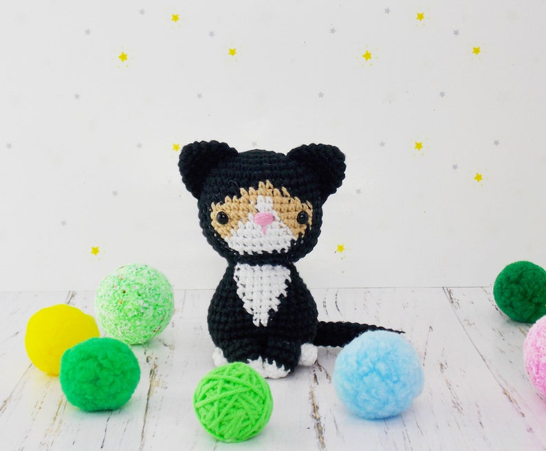 May include: A black and white crocheted cat toy with orange accents. The cat is sitting on a white surface with colorful balls of yarn.