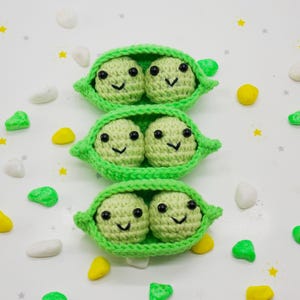 Pu&ograve; includere: Tre baccelli di piselli all'uncinetto, ciascuno con due piselli sorridenti con occhi e bocche neri. I baccelli sono verde brillante e i piselli sono di un verde pi&ugrave; chiaro. Sfondo bianco con elementi decorativi sparsi.