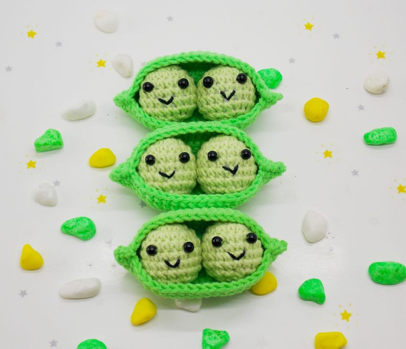 Pu&ograve; includere: Tre baccelli di piselli all'uncinetto, ciascuno con due piselli sorridenti con occhi e bocche neri. I baccelli sono verde brillante e i piselli sono di un verde pi&ugrave; chiaro. Sfondo bianco con elementi decorativi sparsi.