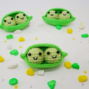 Pu&ograve; includere: Tre baccelli di piselli all'uncinetto, ciascuno con due piselli sorridenti con occhi neri e sorrisi cuciti. I baccelli sono verde brillante e i piselli sono verde pi&ugrave; chiaro. Pietre decorative colorate sono sparse.