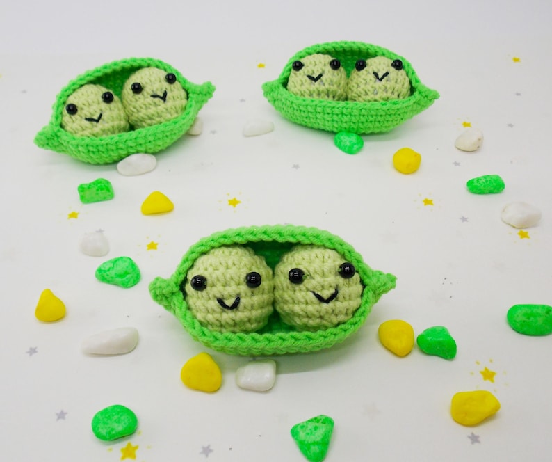 Pu&ograve; includere: Tre baccelli di piselli all'uncinetto, ciascuno con due piselli sorridenti con occhi neri e sorrisi cuciti. I baccelli sono verde brillante e i piselli sono verde pi&ugrave; chiaro. Pietre decorative colorate sono sparse.