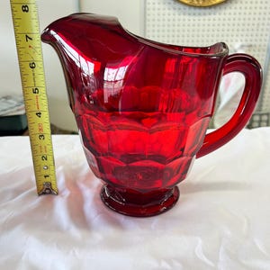 May include: A ruby red glass pitcher with a faceted design. The pitcher has a handle and a spout. The height of the pitcher is approximately 7 inches. The pitcher is sitting on a white surface.