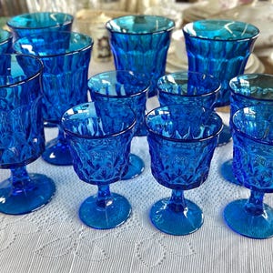 May include: A collection of cobalt blue glassware, including stemmed goblets and tumblers, arranged on a white tablecloth. The glasses have a textured, diamond-like pattern, reflecting light. Other dishware is visible in the background.