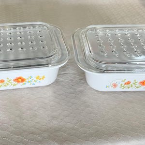 May include: Two white rectangular baking dishes with clear glass lids. The lids have a grid of small holes. Floral designs in orange, yellow, and blue decorate the front of the dishes. The dishes are on a light-colored surface.