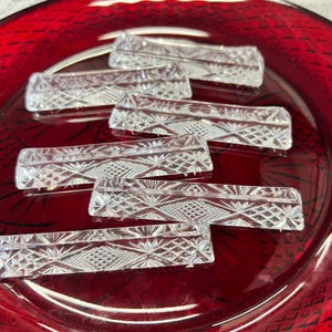 May include: Clear, rectangular crystal knife rests with intricate geometric patterns are arranged on a deep red glass plate. The knife rests are designed to keep knives from touching the table.