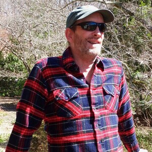 May include: A man wearing a blue and red plaid flannel shirt with a green baseball cap and sunglasses.