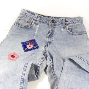 K&ouml;nnte beinhalten: Hellblaue Jeans aus Denim mit zwei gestickten Blumen-Patches. Die Jeans haben einen klassischen F&uuml;nf-Taschen-Stil und einen Knopfverschluss.