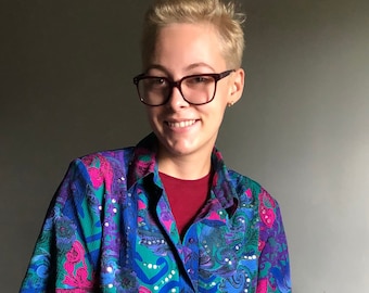 May include: A person wearing a short-sleeved, multi-colored button-down shirt with a floral pattern and rhinestones. The shirt is green, blue, pink, and purple. The person is also wearing blue jeans and a watch.