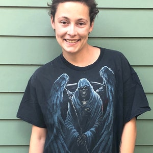 Könnte beinhalten: Ein schwarzes T-Shirt mit einem grafischen Aufdruck einer Kapuzenfigur mit Flügeln und einer Sense. Die Figur sitzt auf einem Schädelhaufen. Das T-Shirt ist überdimensioniert und hat einen Used-Look.