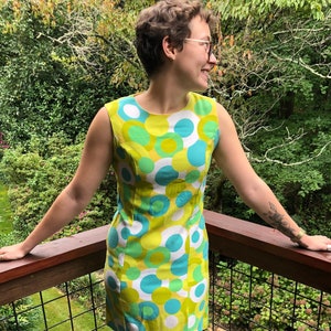 Könnte beinhalten: Eine Frau trägt ein ärmelloses Kleid mit einem geometrischen Muster in Grün, Weiß und Blau. Das Kleid hat einen runden Ausschnitt und eine figurbetonte Silhouette. Sie steht auf einem Holzdeck mit einem Geländer.