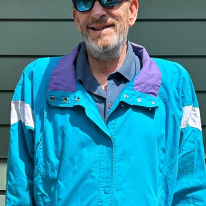 May include: A turquoise jacket with a purple collar and white accents on the sleeves. The jacket has a button-up front and a relaxed fit. The jacket is part of a vintage clothing collection.