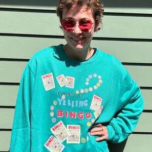 May include: Teal blue sweatshirt with a graphic design of bingo cards and the text "Happiness is yelling bingo".