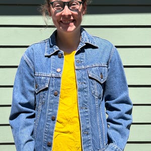 May include: A woman wearing a blue denim jacket over a yellow t-shirt. She is standing in front of a light green wall.