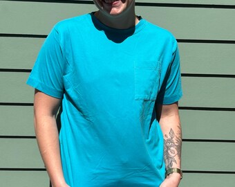 May include: A turquoise blue t-shirt with a single pocket on the left side. The shirt is made of a soft, comfortable fabric.