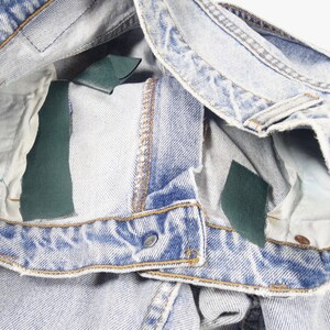 K&ouml;nnte beinhalten: Hellblaue Jeans aus Denim mit einem kleinen rosa Blumenpatch und zwei gr&uuml;nen Stofflabels an der Innenseite des Bunds.