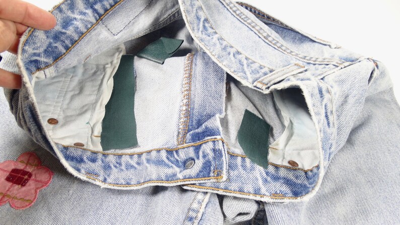 K&ouml;nnte beinhalten: Hellblaue Jeans aus Denim mit einem kleinen rosa Blumenpatch und zwei gr&uuml;nen Stofflabels an der Innenseite des Bunds.