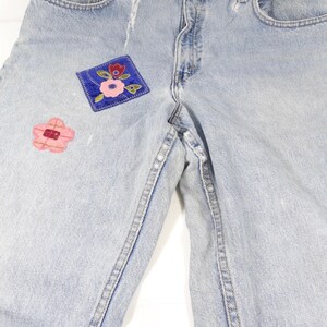 K&ouml;nnte beinhalten: Hellblaue Jeans aus Denim mit zwei gestickten Blumen-Patches. Die Patches sind in Rosa, Rot und Blau.