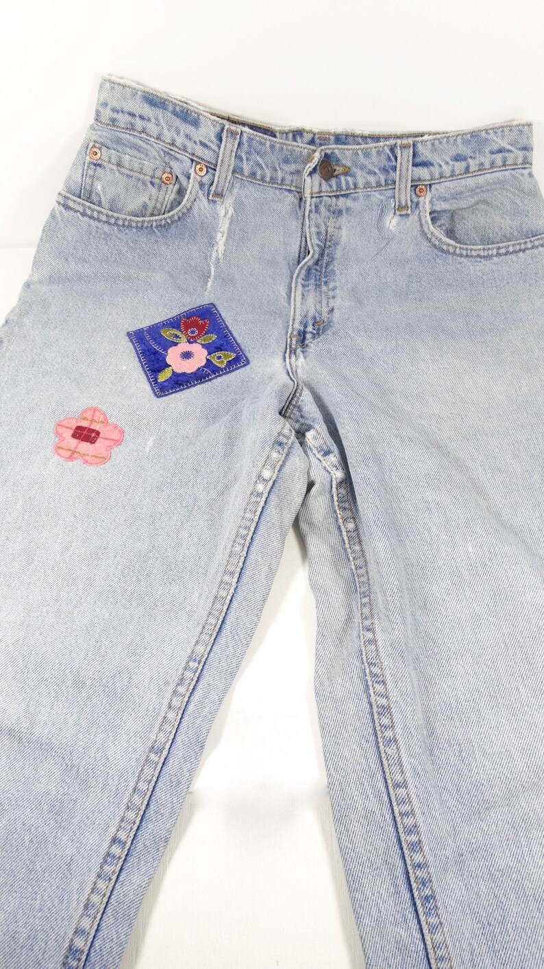 K&ouml;nnte beinhalten: Hellblaue Jeans aus Denim mit zwei gestickten Blumen-Patches. Die Patches sind in Rosa, Rot und Blau.