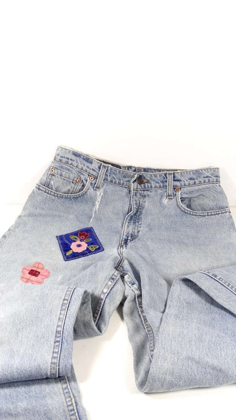 K&ouml;nnte beinhalten: Hellblaue Jeans aus Denim mit zwei gestickten Blumen-Patches. Die Jeans haben einen klassischen F&uuml;nf-Taschen-Stil und einen Knopfverschluss.
