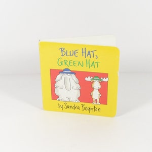 May include: A yellow book with the title "Blue Hat, Green Hat" by Sandra Boynton. The cover features an illustration of an elephant wearing a blue hat and a moose wearing a green hat.