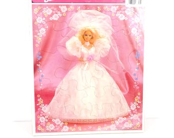 May include: A Barbie frame-tray puzzle depicting a bride doll in a white wedding dress and veil. The puzzle has a pink background with floral accents and the Barbie logo at the top. The puzzle is in a rectangular box.