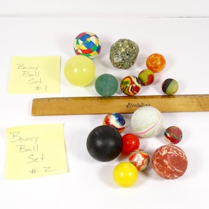 May include: A collection of colorful bouncy balls of various sizes and textures, including a yellow, green, and red ball. A wooden ruler and two yellow sticky notes with handwritten text are also present.
