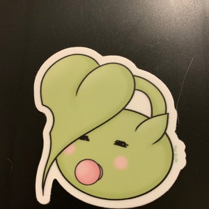 May include: A green cartoon character with a large heart-shaped head and a pink mouth. The character has closed eyes and rosy cheeks.