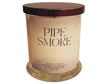 May include: A cylindrical glass candle with a brown lid. The candle has a label with the words "PIPE SMOKE" in a bold font. The label also includes text indicating it is a natural soy candle, hand-poured in Jacksonville, FL, with a 60+ hour burn time and 12 fl oz.