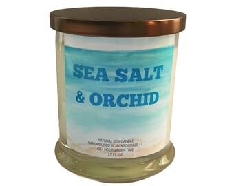 May include: A clear glass candle jar with a bronze lid. The label reads "SEA SALT & ORCHID" over a watercolor beach scene. The candle is a natural soy candle, hand-poured in Jacksonville, FL, with a 60+ hour burn time and 12 fl oz.