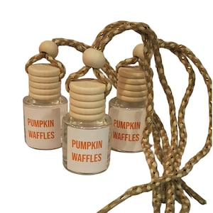 Pode incluir: Três pequenos frascos de vidro transparente com tampas de madeira clara e cordões para pendurar. Cada frasco tem um rótulo branco com o texto PUMPKIN WAFFLES impresso em laranja. Os frascos estão suspensos por cordões trançados.
