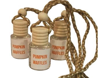 May include: Three small, clear glass bottles with light-colored wooden caps and hanging cords. Each bottle features a white label with the text PUMPKIN WAFFLES printed in orange. The bottles are suspended by braided tan and gold-flecked cords.