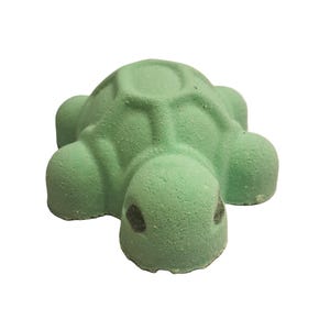 May include: A light green turtle-shaped bath bomb. The bath bomb has a textured surface and a detailed shell design. The turtle's head features two small, dark spots. The bath bomb is designed for use in the bathtub.