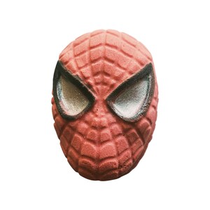 Puede incluir: Una bomba de baño roja con forma de máscara de Spider-Man. La máscara tiene un patrón de telaraña texturizado y ojos grandes de color plateado con contorno negro. La bomba de baño tiene forma esférica.