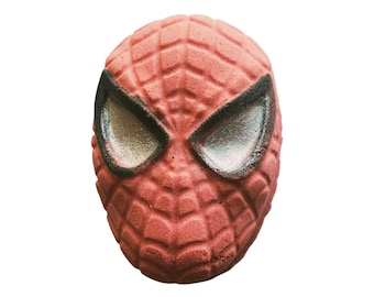 May include: A red bath bomb shaped like Spider-Man's mask. The mask has a textured web pattern and large, silver-colored eyes outlined in black. The bath bomb is a solid, spherical shape.